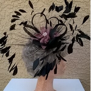 May include: A black feather hat with a large, dark pink flower and a single rhinestone in the center. The hat is on a mannequin head.