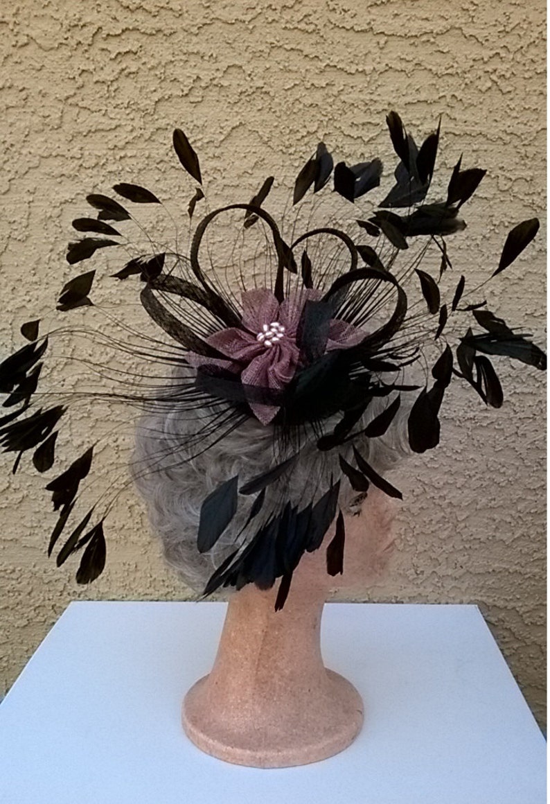 May include: A black feather hat with a large, dark pink flower and a single rhinestone in the center. The hat is on a mannequin head.