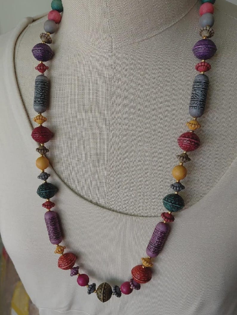 May include: A vibrant beaded necklace with a mix of bead shapes and sizes. The necklace showcases beads in purple, red, green, yellow, and grey hues. Some beads are cylindrical with etched patterns, while others are round or oval.
