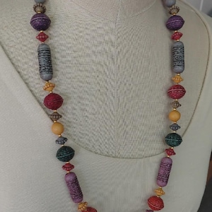 May include: A long, colourful beaded necklace with a variety of shapes and sizes. The necklace features beads in shades of purple, red, green, yellow, and grey. Some beads have intricate patterns, while others are smooth. The necklace is displayed on a white mannequin.