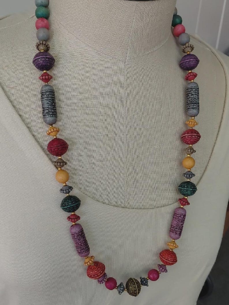 May include: A long, colourful beaded necklace with a variety of shapes and sizes. The necklace features beads in shades of purple, red, green, yellow, and grey. Some beads have intricate patterns, while others are smooth. The necklace is displayed on a white mannequin.