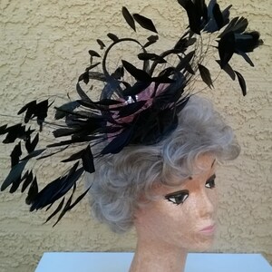 May include: A black feather hat with a pink flower accent. The hat is worn on a mannequin with gray hair.