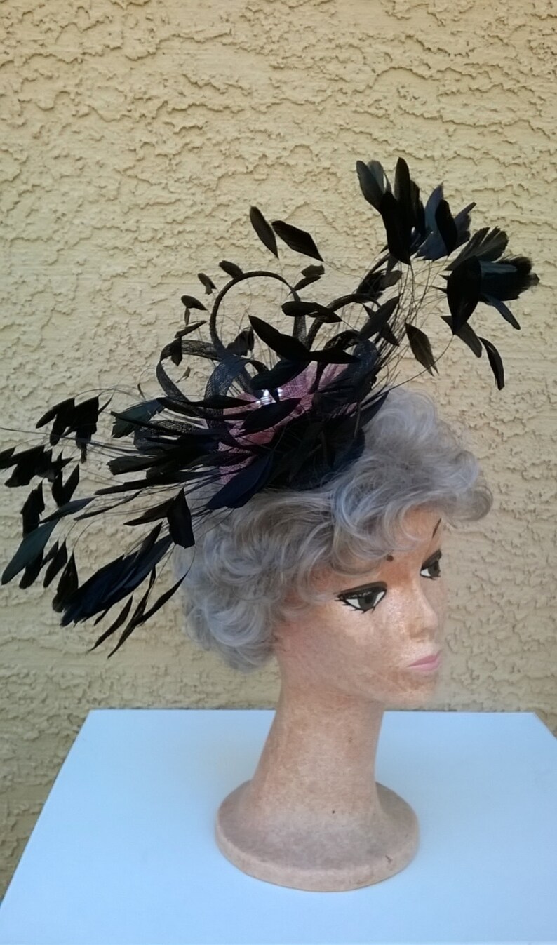 May include: A black feather hat with a pink flower accent. The hat is worn on a mannequin with gray hair.