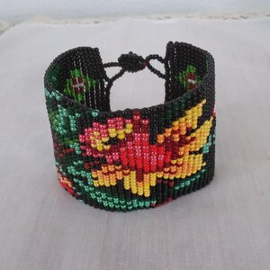 May include: A wide, beaded bracelet with a floral design. The bracelet is primarily black, with a central flower pattern in shades of red, yellow, and orange, accented by green leaves. The bracelet fastens with a beaded loop and knot closure.