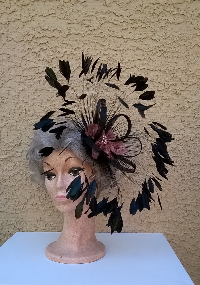 May include: A black feather hat with a large, pink flower accent. The hat is worn on a mannequin head.