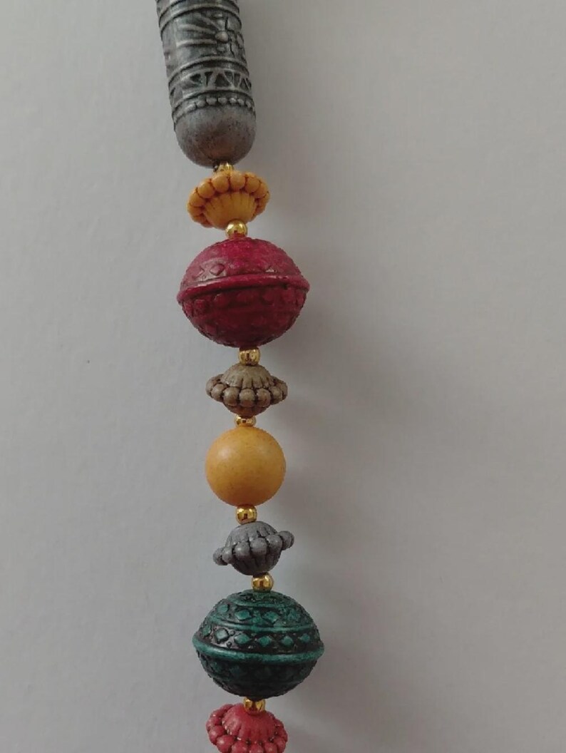 May include: A necklace featuring a variety of beads in red, green, yellow, and grey. The beads have diverse shapes and textures, with gold-coloured accents. The top bead is cylindrical and grey, while the others are spherical or flower-shaped.