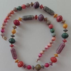 May include: A multi-coloured beaded necklace with a variety of shapes and sizes. The necklace features round, oval, and cylindrical beads in shades of pink, green, yellow, and grey. Some beads have textured patterns and gold-coloured accents. The necklace is displayed on a white background.