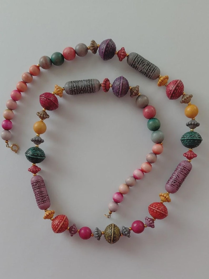 May include: A multi-coloured beaded necklace with a variety of shapes and sizes. The necklace features round, oval, and cylindrical beads in shades of pink, green, yellow, and grey. Some beads have textured patterns and gold-coloured accents. The necklace is displayed on a white background.