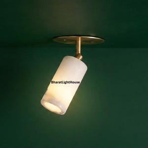 May include: A cylindrical white stone spotlight fixture with a brass-colored metal base and swivel joint. The light is mounted to a dark green surface and features the text BharatLightHouse printed in black on the side of the stone cylinder.