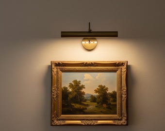 May include: A landscape painting in a gold ornate frame is illuminated by a brass picture light. The painting depicts trees, a river, and a cloudy sky. The light fixture has a circular base and a rectangular light bar.