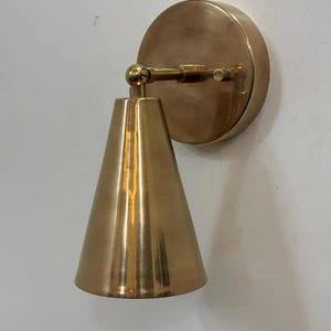 May include: A brass wall sconce with a conical shade and a circular backplate. The sconce is mounted on a white wall. The shade is cone-shaped and the backplate is round. The sconce is made of brass.