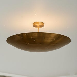 Puede incluir: Una lámpara de techo de color latón con una pantalla poco profunda en forma de cuenco. La pantalla tiene pequeñas perforaciones espaciadas uniformemente. La lámpara está montada sobre un vástago corto y cilíndrico y una base circular.