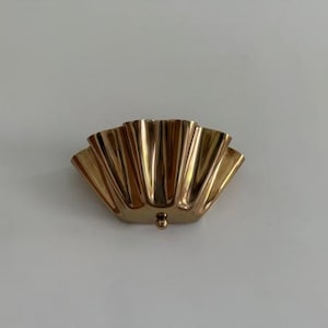 May include: A gold-colored, fan-shaped decorative object. The object has a series of vertical, curved ridges, creating a scalloped appearance. A small, round knob is at the bottom center. The object is likely made of metal.