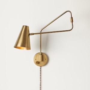 May include: A brass-toned wall sconce with a conical shade and adjustable arm. The sconce features a circular base with a switch and a long, brown electrical cord. The design is minimalist and modern, suitable for various interior styles.