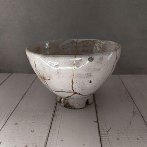 Może przedstawiać: Ręcznie wykonana ceramiczna miska z teksturowanym szkliwem w kolorze złamanej bieli i nieregularnym, organicznym kształcie. Miska posiada widoczne pęknięcia naprawione złotym lakierem, tworząc na powierzchni charakterystyczny wzór przypominający żyłki.
