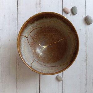 Może przedstawiać: Okrągła ceramiczna miska z gradientem brązowych i kremowych odcieni z widocznymi pęknięciami naprawionymi za pomocą złotego lakieru. Wewnętrzna powierzchnia ma teksturowane wykończenie, a złote linie tworzą rozgałęziony wzór na misce.