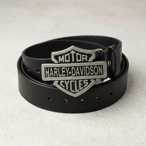 Könnte beinhalten: Ein schwarzer Ledergürtel mit einer silberfarbenen Harley-Davidson-Schnalle. Die Schnalle zeigt das Harley-Davidson-Logo mit den Worten "MOTOR", "HARLEY-DAVIDSON" und "CYCLES". Der Gürtel ist aufgerollt und hat mehrere Löcher zur Größenanpassung.