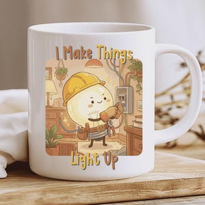 Op de afbeelding: Witte keramische mok met een cartoon illustratie van een gloeilamp karakter met een gele helm die aan een elektrisch paneel werkt. De tekst "I Make Things Light Up" staat op de mok.