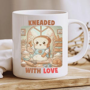 Puede incluir: Taza de cerámica blanca con una ilustración colorida de un personaje de rebanada de pan amasando masa. La taza tiene el texto "KNEADED WITH LOVE" en una fuente juguetona. El fondo muestra una escena de panadería con pan y un gato.