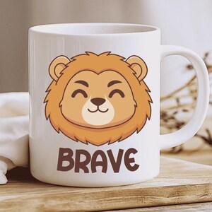 Op de afbeelding: Witte keramische mok met een cartoon leeuwenkop met een glimlach en een bruine manen. Het woord "BRAVE" is in bruine blokletters onder de leeuwenkop gedrukt. De mok heeft een wit handvat.