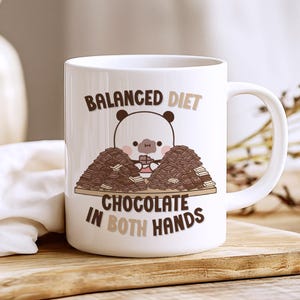Puede incluir: Taza de cerámica blanca con una ilustración de un panda de dibujos animados. El panda está rodeado de barras de chocolate y el texto dice "BALANCED DIET CHOCOLATE IN BOTH HANDS". La taza tiene un asa blanca y está sobre una superficie de madera.