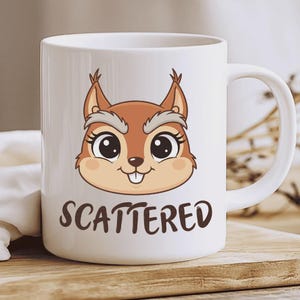 Puede incluir: Taza de cerámica blanca con la cara de una ardilla de dibujos animados y la palabra "SCATTERED". La ardilla tiene pelaje marrón y una cara marrón claro. La taza tiene un asa blanca. La taza está sobre una superficie de madera.