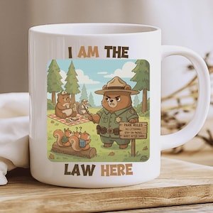 Puede incluir: Taza de cerámica blanca con una ilustración de dibujos animados de un oso guardabosques y el texto "I AM THE LAW HERE". El diseño incluye otros osos y ardillas en un bosque, con un letrero que dice "Park Rules".
