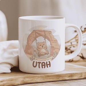 Könnte beinhalten: Weiße Keramiktasse mit einer Cartoon-Illustration eines Bären, der unter einem Felsbogen in einer Wüstenlandschaft schaukelt. Das Wort "UTAH" ist in Braun aufgedruckt. Die Tasse hat einen gebogenen Henkel.