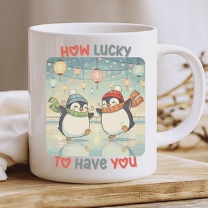 Op de afbeelding: Witte keramische mok met een illustratie van twee pinguïns die schaatsen. De pinguïns dragen mutsen en sjaals, met de tekst "How Lucky To Have You" in roze en rood. De achtergrond toont lantaarns en een besneeuwde scène.