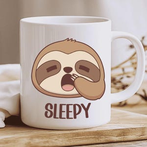 Puede incluir: Taza de cerámica blanca con un perezoso de dibujos animados bostezando y cubriéndose la boca con la pata. El perezoso es marrón y beige con los ojos cerrados. La palabra "SLEEPY" está impresa en marrón debajo.
