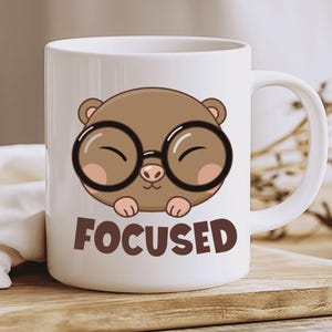 Puede incluir: Taza de cerámica blanca con un dibujo de una nutria de dibujos animados con gafas negras grandes. La nutria es marrón con mejillas rosadas y está sonriendo. La palabra "FOCUSED" está impresa en letras marrones.