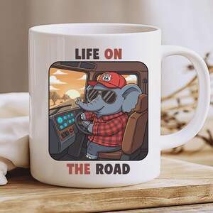 Op de afbeelding: Witte keramische mok met een illustratie van een olifant die een vrachtwagen bestuurt. De olifant draagt een zonnebril, een Route 66-pet en een rood geruit overhemd. De mok heeft de tekst "LIFE ON THE ROAD" in rood en bruin.