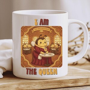 Puede incluir: Taza de cerámica blanca con una abeja de dibujos animados con un vestido rojo, sentada a una mesa en un restaurante. La taza tiene el texto "I AM THE QUEEN" y "THE HIVE RESTAURANT". El fondo tiene un diseño de panal.