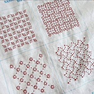 May include: A white fabric sampler featuring four distinct sashiko embroidery patterns stitched in dark red thread. Each square section displays a unique geometric design, including floral and interlocking grid motifs, with thin blue guide lines.