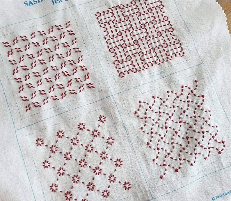 May include: A white fabric sampler featuring four distinct sashiko embroidery patterns stitched in dark red thread. Each square section displays a unique geometric design, including floral and interlocking grid motifs, with thin blue guide lines.
