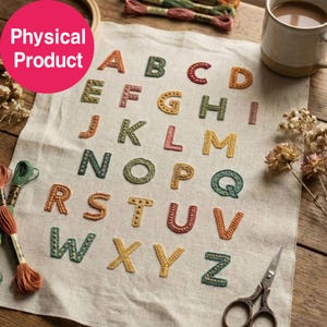 Puede incluir: Una pieza de tela de color claro con el alfabeto bordado en varios colores. Cada letra está cosida con un patrón texturizado. La tela se muestra junto a hilo de bordar, una taza y un par de tijeras de metal.
