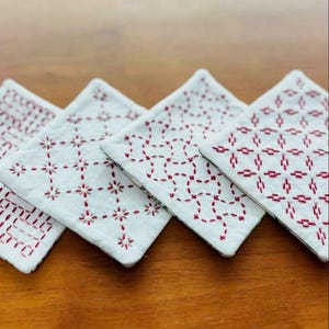 May include: Four square fabric coasters featuring white cotton surfaces with intricate red thread embroidery in various geometric patterns. Each coaster displays a unique stitched design, showcasing detailed needlework and clean white fabric edges.