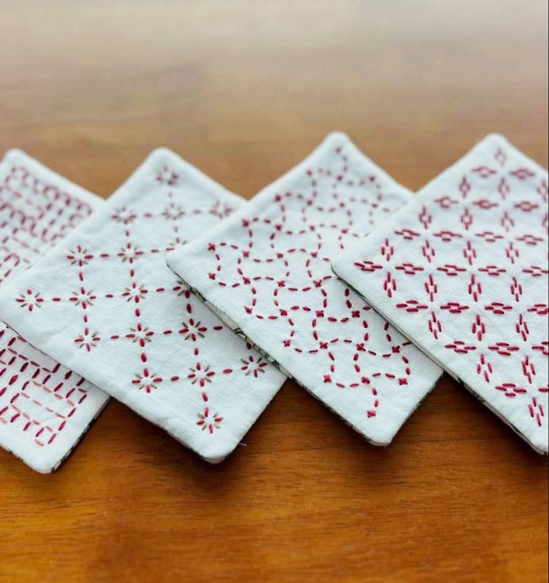 May include: Four square fabric coasters featuring white cotton surfaces with intricate red thread embroidery in various geometric patterns. Each coaster displays a unique stitched design, showcasing detailed needlework and clean white fabric edges.
