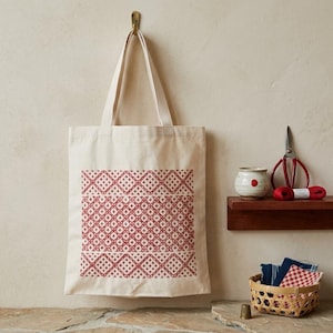 May include: A cream-colored canvas tote bag featuring a repeating geometric pattern in deep red ink. The design consists of interconnected diamond shapes and small crosses. The bag has two long shoulder straps and hangs from a wall hook.