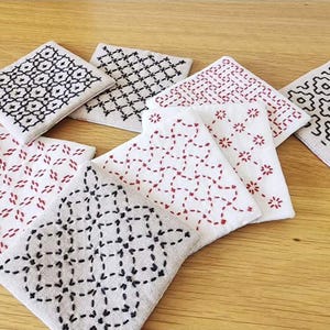 May include: A collection of square fabric coasters featuring intricate hand-stitched embroidery patterns. The designs include geometric shapes and floral motifs in black and red thread on white and light gray linen-like fabric backgrounds.