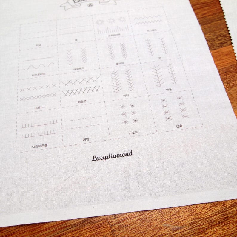 May include: A white fabric with a grid of various stitch patterns, including straight, zigzag, and decorative stitches. The text "Lucydiamond" is printed in black script at the bottom of the fabric. The fabric is on a wooden surface.
