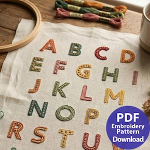 Könnte beinhalten: Gesticktes Alphabet-Muster mit farbigem Garn. Die Buchstaben A bis U sind in verschiedenen Farben gestickt, darunter Orange, Grün, Rosa und Gelb. Stickgarn und ein Holzrahmen sind sichtbar. Ein blauer Kreis mit Text lautet "PDF Embroidery Pattern Download."