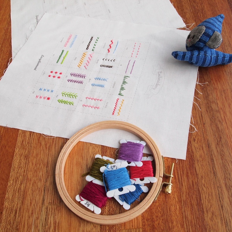 May include: A wooden embroidery hoop with colorful thread spools, a fabric with various stitch examples, and a small blue stuffed animal on a wooden surface. The fabric has the word "Embroidery" printed on it.
