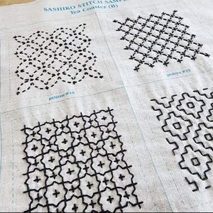May include: A beige fabric sampler featuring four distinct black Sashiko embroidery patterns. The text SASHIKO STITCH SAMPLE Tea Coaster (B) is printed at the top, with labels pattern #18 and pattern #19 visible below the individual stitched squares.