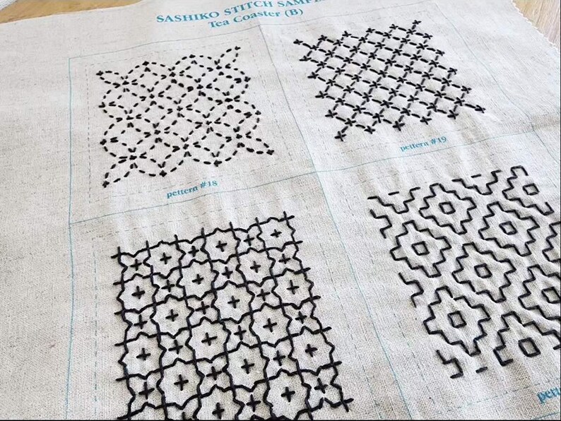May include: A beige fabric sampler featuring four distinct black Sashiko embroidery patterns. The text SASHIKO STITCH SAMPLE Tea Coaster (B) is printed at the top, with labels pattern #18 and pattern #19 visible below the individual stitched squares.