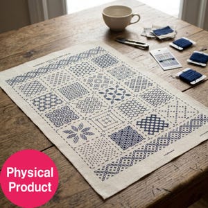 Könnte beinhalten: Ein rechteckiger beigefarbener Stoff-Sampler mit einem Raster aus verschiedenen dunkelblauen Sashiko-Stickmustern. Der Text "SASHIKO STITCH SAMPLER 20" ist oben aufgedruckt. Der Stoff wird von mehreren blauen Garnrollen und Nadeln begleitet.