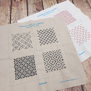 May include: Two rectangular fabric panels featuring Sashiko stitch sampler patterns. The top panel is white with red stitching, while the bottom panel is beige with black stitching. Both include blue printed text labels and grid outlines.