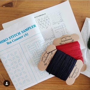 May include: A Sashiko stitch sampler tea coaster kit featuring white fabric with blue printed patterns, a printed instruction sheet, and two cardboard spools wrapped with red and black embroidery thread labeled Lucydiamond Custom Clothing.