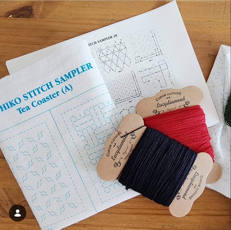 May include: A Sashiko stitch sampler tea coaster kit featuring white fabric with blue printed patterns, a printed instruction sheet, and two cardboard spools wrapped with red and black embroidery thread labeled Lucydiamond Custom Clothing.