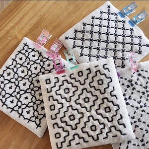 May include: Four square pieces of light beige fabric featuring black hand-stitched embroidery patterns. Each fabric square is secured with colorful plastic sewing clips in shades of pink, blue, and green, displaying geometric designs.